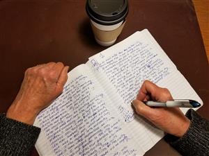The hands of a writer working in a notebook. A cup of coffee in the backgroud. 